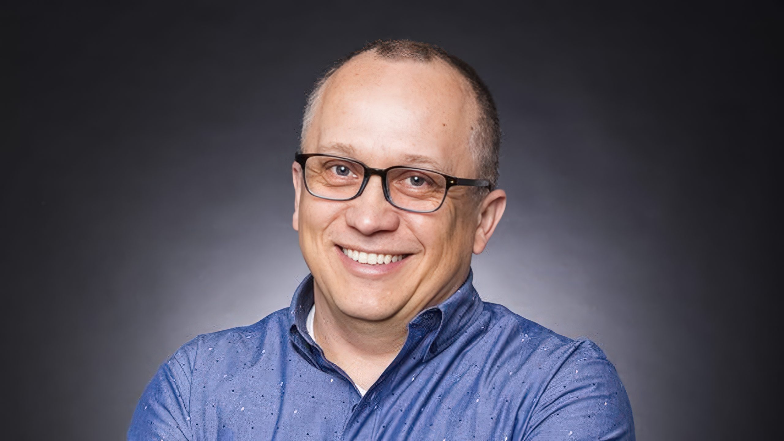 A person wearing glasses and a blue patterned shirt, smiling against a dark gray background.