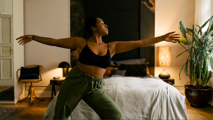Μια γυναίκα με μαύρο αθλητικό σουτιέν και πράσινο παντελόνι που απλώνεται στην άκρη του κρεβατιού σε δωμάτιο με χαμηλό φωτισμό με φυτό