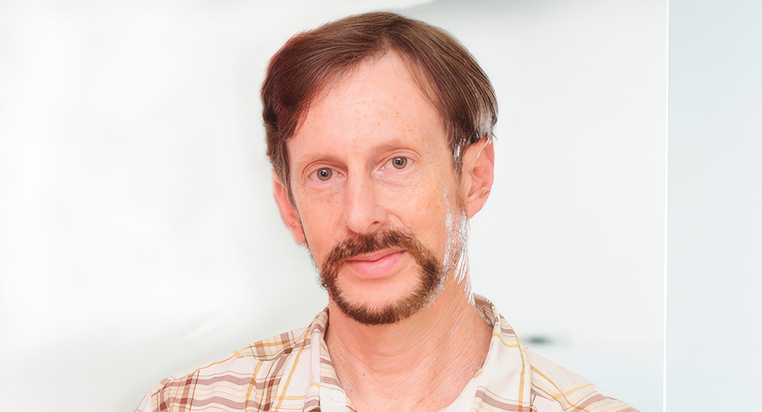 A person with short brown hair and a beard wearing a plaid shirt against a plain background.
