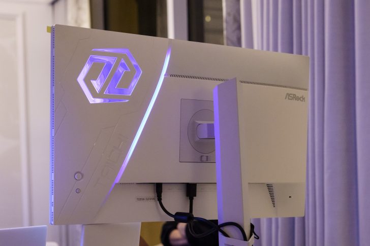 A white ASRock gaming monitor with a glowing purple logo is shown from the back, highlighting its design and connectivity ports.