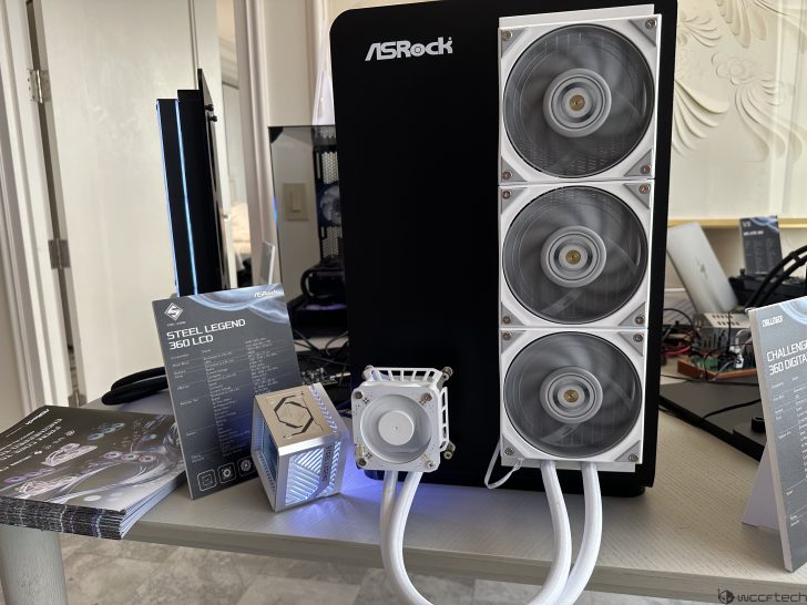An ASRock Steel Legend 360 LCD cooling system is showcased with three fans attached to a black ASRock case, accompanied by informational pamphlets on the table.
