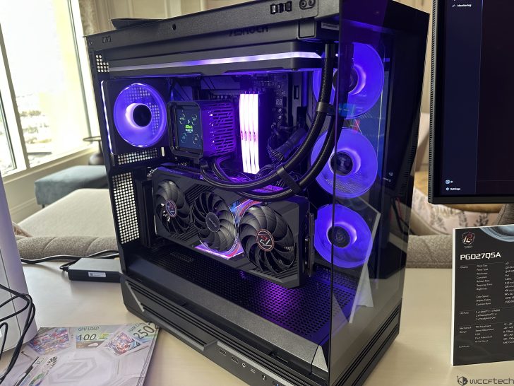 A gaming PC with an NZXT graphics card and liquid cooling system is illuminated with purple LED lights, next to a monitor displaying 'PG027Q5A,' and a pamphlet showing 'GPU LOAD 100%.'.
