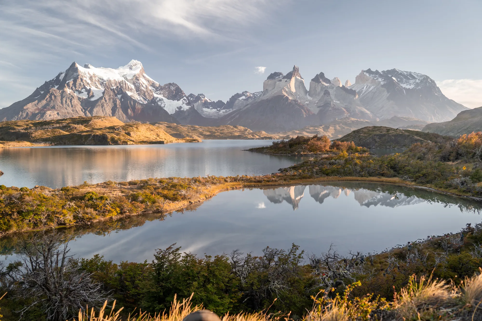 Calm waters act as a perfect mirror, doubling the magnitude of the snow-capped peaks in the distance. The golden hue of the shoreline vegetation emphasises the stillness of a windless autumn morning.