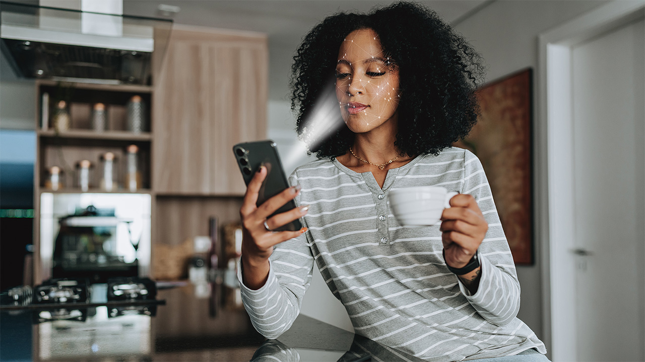 Το άτομο, καθόταν σε μια κουζίνα κρατώντας μια κούπα, σαρώνοντας το πρόσωπό του με το smartphone του