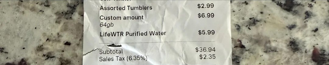 Μια απόδειξη σε μια μαρμάρινη επιφάνεια αναφέρει αντικείμενα όπως «Ποικίλα ποτηράκια» για 2,99 $, «Προσαρμοσμένο ποσό 64 gb» για 6,99 $ και «LifeWTR Purified Water» για 5,99 $, με υποσύνολο 36,94 $ και φόρο επί των πωλήσεων 2,35 $.