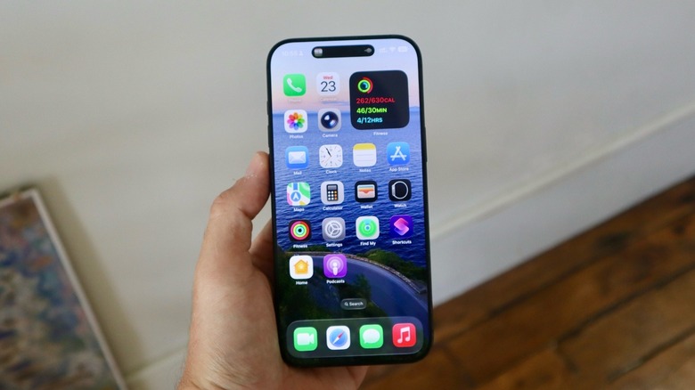 Ένα χέρι που κρατά ένα iPhone 16 Pro Max. Υπάρχει μισό πλαίσιο στο βάθος κοντά σε έναν τοίχο.