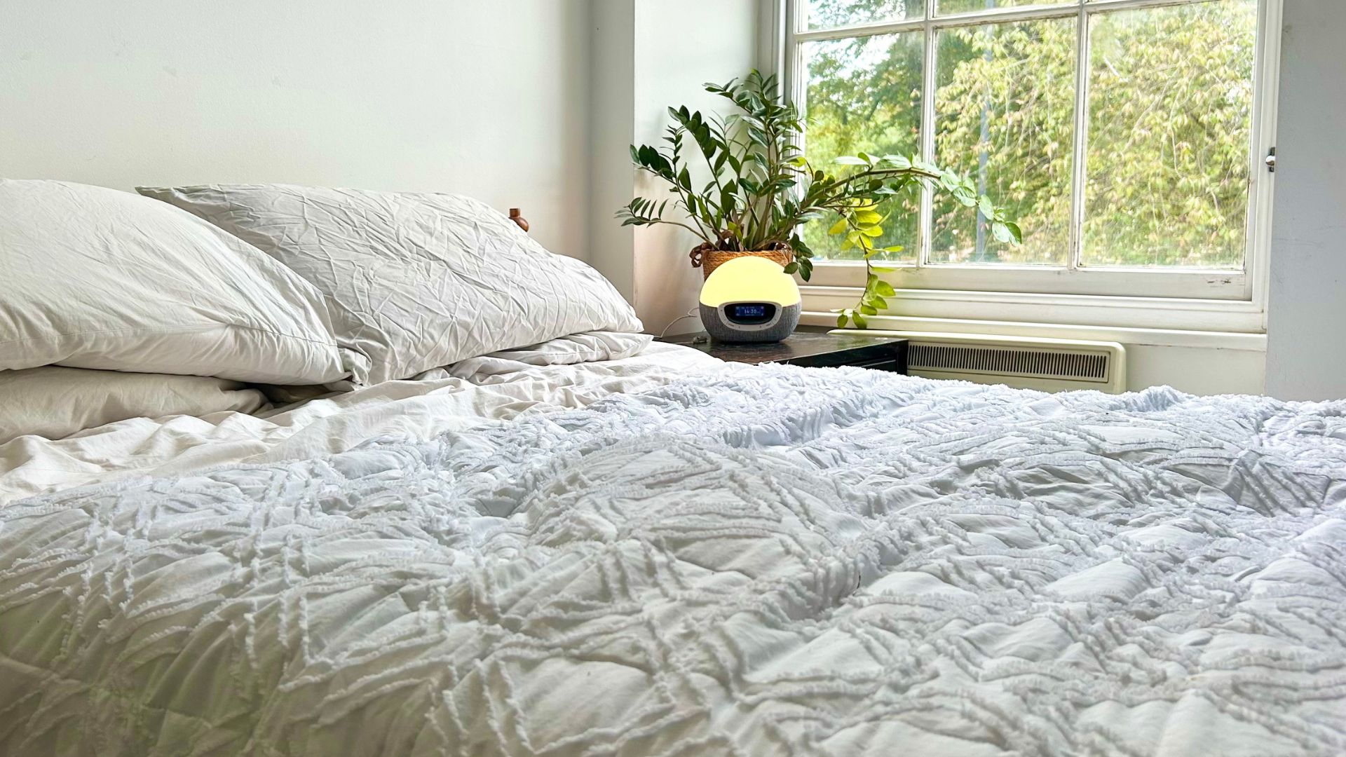 Το φως Lumie Bodyclock Shine 300 στο κομοδίνο δίπλα σε ένα κρεβάτι μπροστά από ένα ανοιχτό παράθυρο.