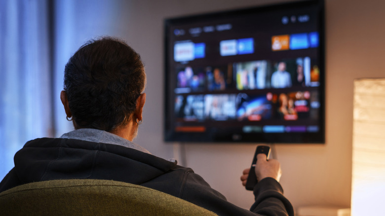 Άνδρας που χρησιμοποιεί ένα τηλεχειριστήριο υπερύθρων για να επιλέξει από τις επιλογές ροής σε μια επιτοίχια έξυπνη τηλεόραση