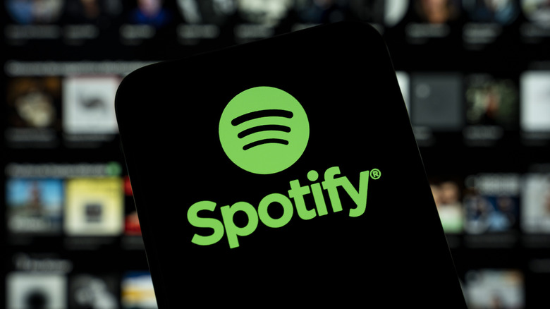 Ένα smartphone που εμφανίζει το λογότυπο Spotify, με ασαφές υπόβαθρο που φαίνεται να είναι οθόνη επιλογής άλμπουμ από το Spotify